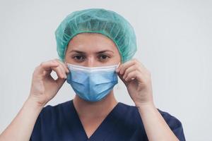 dichtbij omhoog van vrouw dokter of wetenschapper met een medisch masker en chirurgisch pet over- grijs achtergrond. ze is aanpassen masker met foto