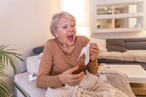 verkoudheid en griep. portret van ziek senior vrouw gevangen koud, gevoel ziek en niezen in papier veeg. ongezond ouderling vrouw gedekt in deken afvegen neus. gezondheidszorg concept. foto