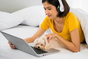 Aziatisch vrouw gebruik makend van een laptop computer en aan het liegen Aan een bed met shihtzu hond foto