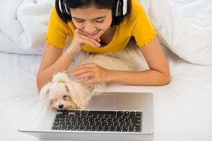 gelukkig vrouw gebruik makend van een laptop computer en aan het liegen Aan een bed met shihtzu hond foto