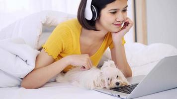 Aziatisch vrouw gebruik makend van een laptop computer en aan het liegen Aan een bed met shihtzu hond foto