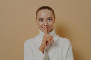 hou het geheim. Jonge aantrekkelijke glimlachende dame die zegt wees stil met de vinger op de lippen, geïsoleerd op beige foto