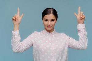 horizontaal schot van zelfverzekerde brunette jonge dame in formeel overhemd wijst naar boven met beide voorvingers, toont richting hierboven, donker gekamd haar, geïsoleerd op blauwe achtergrond. advertentie concept foto