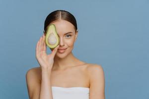 gezichtsverzorging concept. charmante tedere vrouw met gekamd donker haar heeft gloeiende huid bedekt oog met de helft van avocado krijgt essentiële oliën voor lichaamsbehandeling gewikkeld in badhanddoek geïsoleerd op blauwe muur foto