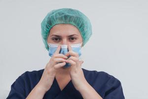 dichtbij omhoog van vrouw dokter of wetenschapper met een medisch masker en chirurgisch pet over- grijs achtergrond. ze is aanpassen masker met foto