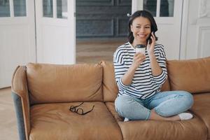 gelukkig meisje maakt telefoon telefoongesprek, zittend Aan sofa met koffie kop Bij huis. aangenaam mobiel gesprek foto