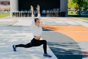 flexibele pasvorm sportvrouw doet sportoefeningen buitenshuis houdt de armen omhoog heeft een blije uitdrukking draagt hoodies leggings en trainers poses op stadion in stedelijke plaats luistert naar favoriete audiotrack. foto