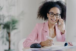 gelukkige afro-amerikaanse vrouw met krullend kapsel, schrijft wat informatie, spreekt via mobiele telefoon, zit aan het bureau tegen een wazige kantoorachtergrond. mensen, technologie, communicatie, bedrijfsconcept foto