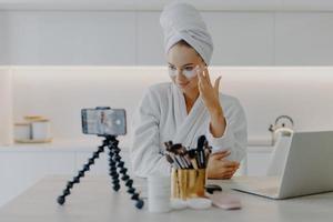 mooi jong vrouwelijk model adverteert voor internetweergaven cosmetisch product geeft om de huid onder de ogen past hydrogelpleisters toe neemt video op smartphone-apparaat op voor haar blog. vloggen concept foto