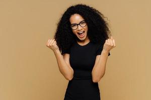 triomfantelijke mooie vrouw met donkere huid heft gebalde vuisten op, schreeuwt van vreugde, viert overwinning, draagt zwarte kleding, staat tegen bruine achtergrond, voelt als winnaar. lichaamstaal, succesconcept foto