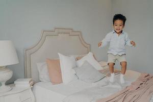 gelukkig zorgeloos gemengd ras etniciteit jongen in vrijetijdskleding die plezier heeft en op bed springt foto