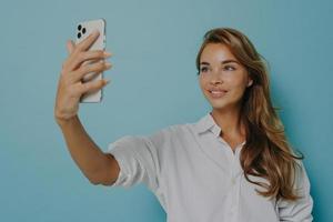 jonge elegante Europese vrouw poseren voor selfie, staande met smartphone in de hand tegen blauwe muur foto