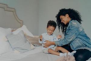 liefhebbende Afro-Amerikaanse moeder die zoon kietelt terwijl ze samen plezier heeft in de slaapkamer foto