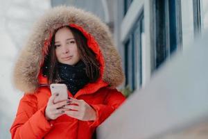 foto van aantrekkelijke vrouw houdt moderne moderne gadget vast, draagt rode jas met capuchon, controleert e-mailbox, verbonden met draadloos internet, staat buiten, geniet van vrije tijd en ijzig winterweer