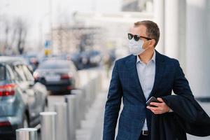 doordachte ondernemer in formele kleding, zonnebril, beschermt zichzelf met steriel medisch masker tegen coronavirusziekte, loopt buiten, houdt mobiele telefoon vast, denkt aan veiligheid. verantwoordelijk gedrag foto