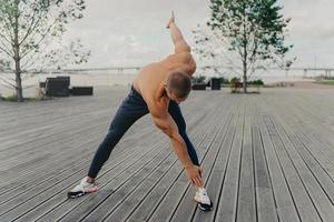 knappe gemotiveerde ongeschoren sportman doet oefeningen op de promenade, warmt op voordat hij gaat rennen, gekleed in actieve kleding, poseert buiten. mannelijke hardloper met naakte torso blijft fit, leidt een gezonde levensstijl foto