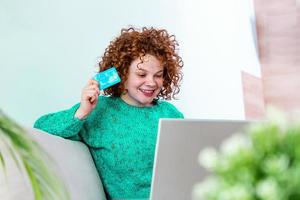 gelukkig jong vrouw Aan sofa boodschappen doen online met debiteren kaart. mooi meisje gebruik makend van laptop computer voor online boodschappen doen Bij huis verliet voor vinden item Aan uitverkoop foto