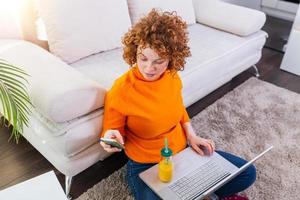 schattig vrouw rood haar- freelancer lezing tekst berichten terwijl zittend Aan verdieping met Open computer in modern huis, jong creatief vrouw werk Aan laptop terwijl hebben oranje sap foto