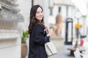 portret van een mooie langharige aziatische vrouw in een zwarte jas, met een wandeltas, glimlachend in een goed humeur in de stad buiten. foto