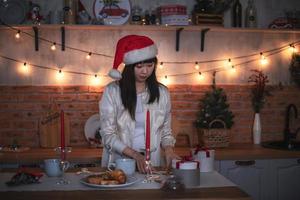 een jong Koreaans vrouw in een de kerstman claus hoed kookt in de avond in een keuken versierd voor kerstmis. foto