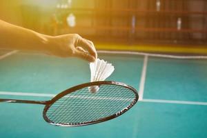 badminton racket en oud wit shuttle Holding in handen van speler terwijl portie het over- de netto vooruit, vervagen badminton rechtbank achtergrond en selectief focus foto