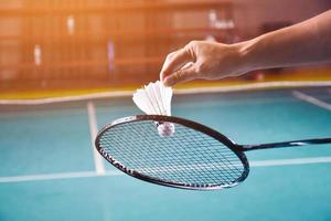 badminton racket en oud wit shuttle Holding in handen van speler terwijl portie het over- de netto vooruit, vervagen badminton rechtbank achtergrond en selectief focus foto