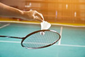 badminton racket en oud wit shuttle Holding in handen van speler terwijl portie het over- de netto vooruit, vervagen badminton rechtbank achtergrond en selectief focus foto