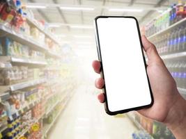 slim boodschappen doen concept mobiel telefoon in hand- in voorkant van goederen schappen in supermarkt en kruidenier op te slaan. blanco wit scherm mockup voor uw eigen creativiteit. foto