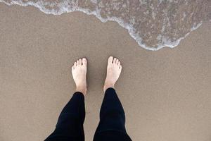 Mens vervelend een wetsuit staand Aan een schoon zanderig strand Aan meer Michigan aan het kijken de water komen naar zijn voeten foto