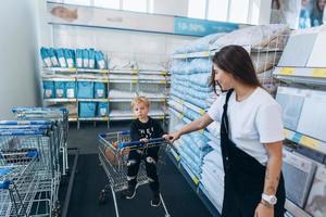 mooi moeder draagt haar weinig zoon in de supermarkt trolley foto