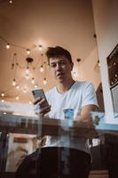 jong, knap Mens toepassingen een smartphone in een cafe. foto achter glas