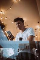 jong, knap Mens toepassingen een smartphone in een cafe. foto achter glas
