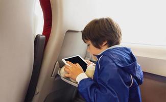 kind op reis door de trein, Aziatisch jongen aan het kijken tekenfilm Aan tablet zittend door de venster. kind in een hoog snelheid uitdrukken trein Aan familie vakantie, vermaak voor jong passagier. foto