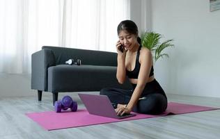 Aziatische sportieve vrouw in sportkleding die aan het trainen is en laptop gebruikt en telefoon thuis in de woonkamer belt, zittend op de vloer met halters op yogamat. sport- en online trainingsconcept foto