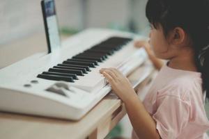 Aziatisch schattig meisje aan het leren online piano muziek- met leraar door mobiel telefoon of tablet. de idee van activiteiten voor de kind Bij huis gedurende quarantaine. muziek- aan het leren studie online concept. foto