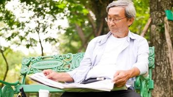 gelukkig Aziatisch Mens zittend Aan een bank en lezing een krant- Bij de park. foto