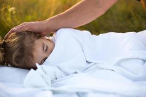 meisje slaapt Aan wit bed in de gras, vers lucht. van vader hand- voorzichtig klopt zijn hoofd. zorg, bescherming, Internationale kinderen dag, mug bijt foto