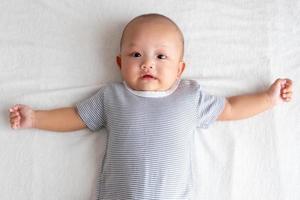 geschokt baby jongen vervelend een gestreept overhemd met een handvol van handen Aan een wit matras foto