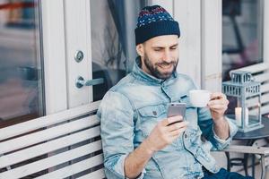 opgetogen gelukkige man met dikke baard en snor, leest positief nieuws op mobiele telefoon, geniet van recreatietijd, drinkt thee in de coffeeshop, heeft een gelukkige uitdrukking. lachende knappe man gebruikt mobiel foto