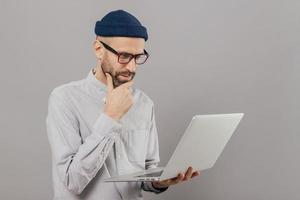 attente student houdt kin vast, gefocust op monitor van laptopcomputer, zoekt informatie voor project, verbonden met draadloos internet, geïsoleerd over grijze studiomuur. netwerken en mensen concept foto