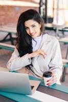 jonge vrouw met donker haar met heldere ogen, volle lippen en een gezonde huid met een witte jas die in café rust en op internet surft met behulp van een laptopcomputer die smakelijke koffie drinkt. schoonheid en jeugdconcept foto