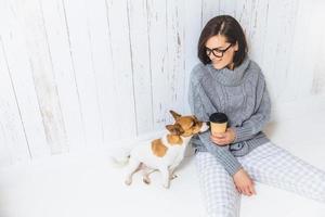 mensen, levensstijl en vrije tijd concept. zorgeloze mooie jonge vrouw gekleed in huishoudelijke kleding, drinkt afhaalkoffie, brengt vrije tijd door met hond, geniet van een rustige sfeer, goed humeur foto