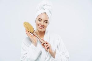 mooie blije vrouw met een gezonde huid en een brede glimlach houdt een houten massageborstel voor het lichaam, draagt een badjas en een gewikkelde zachte handdoek op het hoofd, staat tegen een witte achtergrond. schoonheid, zorgconcept foto