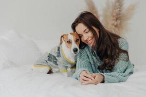 foto van mooie brunette vrouw in gebreide trui ligt samen met hond op zacht bed, brengt graag tijd door met favoriete huisdier, geeft om dieren, blijft thuis tijdens coronavirus quarantaine