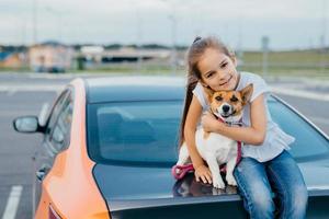 klein aantrekkelijk vrouwelijk kind omhelst haar favoriete hond, zit samen in de kofferbak van de auto, rust uit na een wandeling, geniet van de zomerdag, heeft een vriendschappelijke relatie. kinderen, huisdieren en lifestyle concept. foto