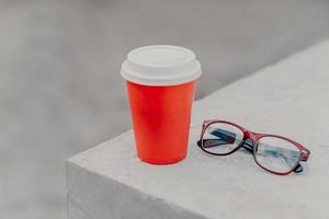 rode wegwerpbeker warme drank op witte betonnen muur, optische bril in de buurt. afhaalkoffie in papieren container. geen mensen. aromatische espresso. foto