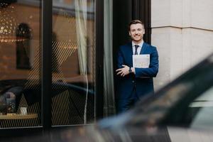 buitenshuis schot van knap mannetje in formeel kleren, staat gekruiste handen, houdt logboek, staat in de buurt cafe, wacht voor collega's, heeft tevreden uitdrukking, teder glimlach Aan gezicht. mensen en levensstijl foto