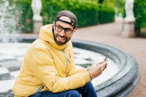 zijwaarts buitenportret van een vrolijke, bebaarde stijlvolle man in een gele jas en een bril die in de buurt van de fontein in het park zit en de moderne decive in handen houdt en gelukkig opzij kijkt terwijl hij de schoonheid van de natuur bewondert. foto