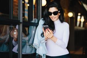 stijlvolle vrouw met donker golvend haar, gekleed in een witte blouse met een jas met een grote zonnebril die een smartphone vasthoudt die online met haar vriendje communiceert. brunette vrouw die nieuwe applicaties bijwerkt foto