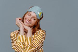 portret van knappe vrolijke vrouw kantelt hoofd en glimlacht zachtjes, verbeeldt iets aangenaams, draagt slaapmasker of blinddoek en pyjama, geïsoleerd over grijze achtergrond, poses in gezellige slaapkamer foto